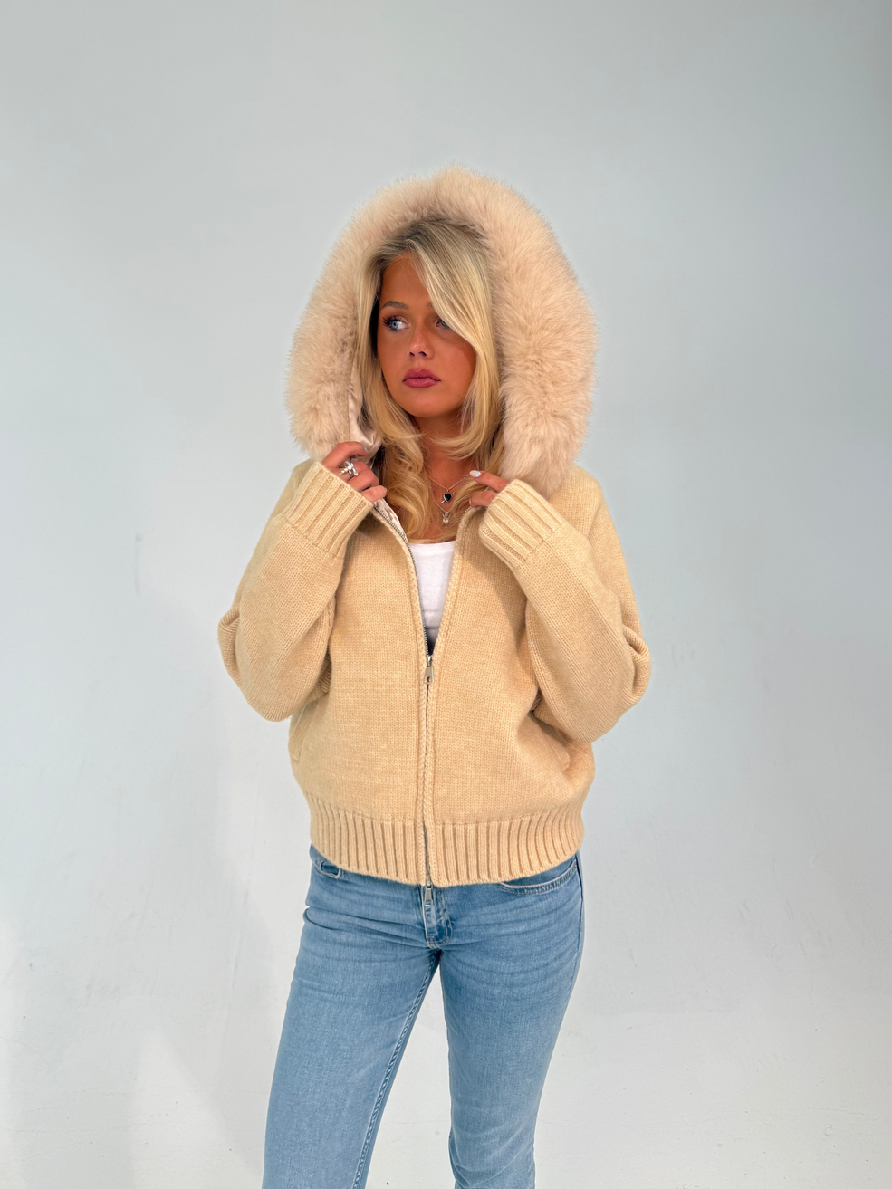 Woman wearing a beige cardigan with a fur-lined hood against a plain background